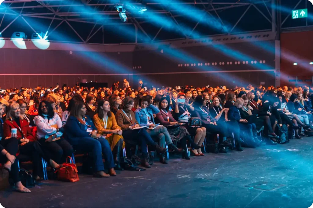 A large conference audience seated in rows in a convention hall, wearing lanyards and holding notebooks and phones, with blue stage lights beaming across the ceiling.