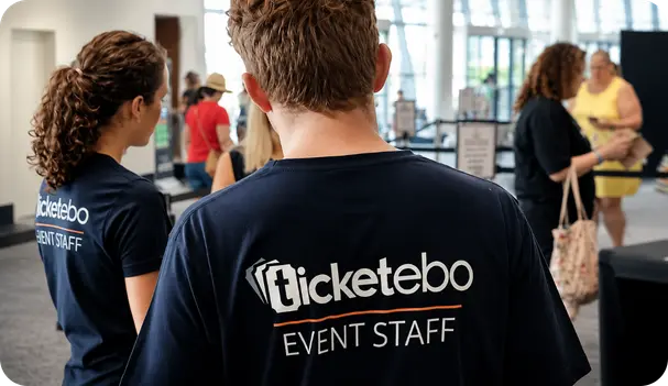 Ticketebo event staff in navy "Event Staff" t-shirts welcoming attendees at the entrance of a bright, glass-walled venue foyer.